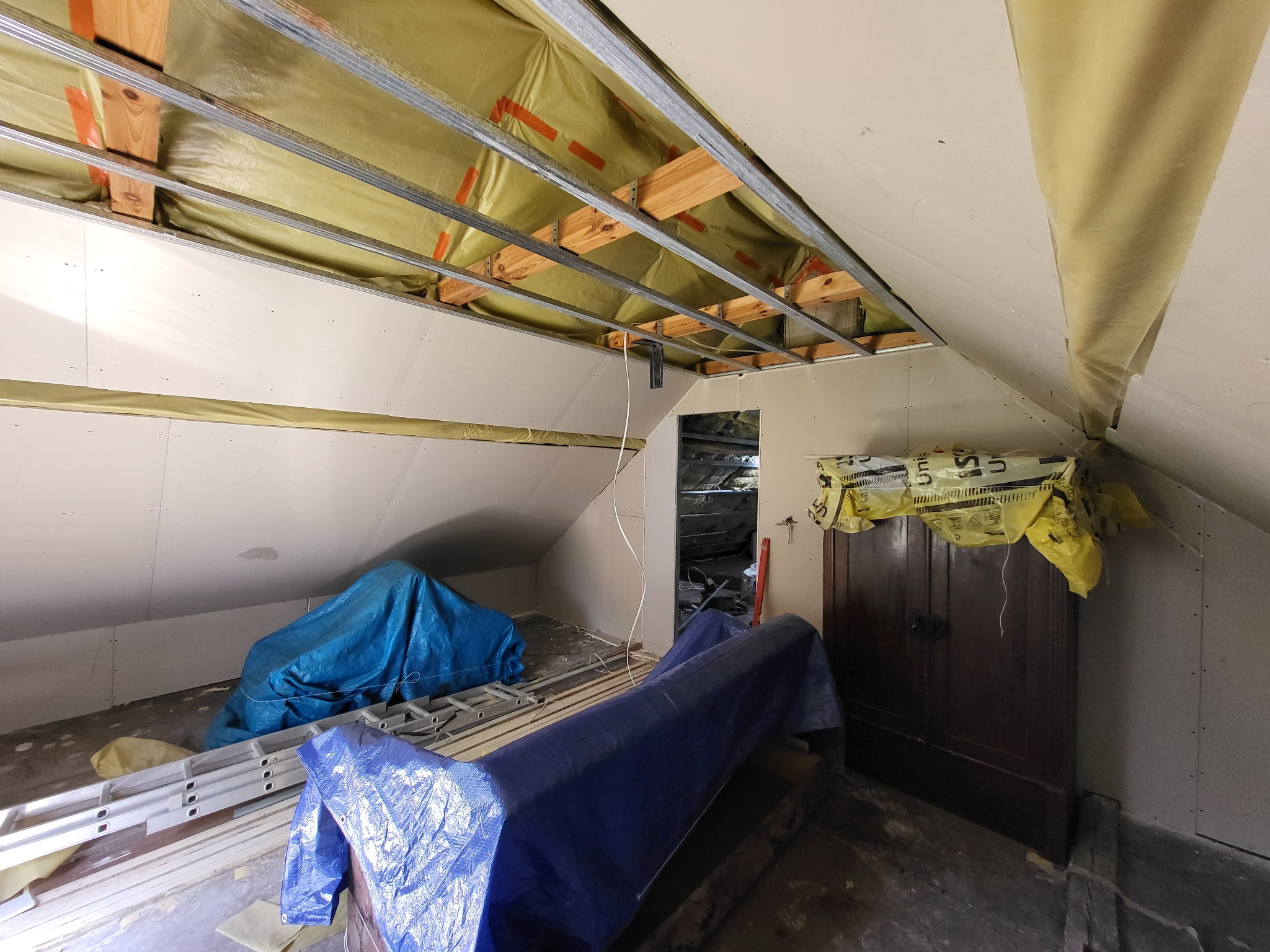 Poddasze w trakcie adaptacji: widoczne metalowe profile sufitowe, częściowo zamontowana płyta gipsowa, izolacja i zabezpieczone meble. Prace wykończeniowe w toku.