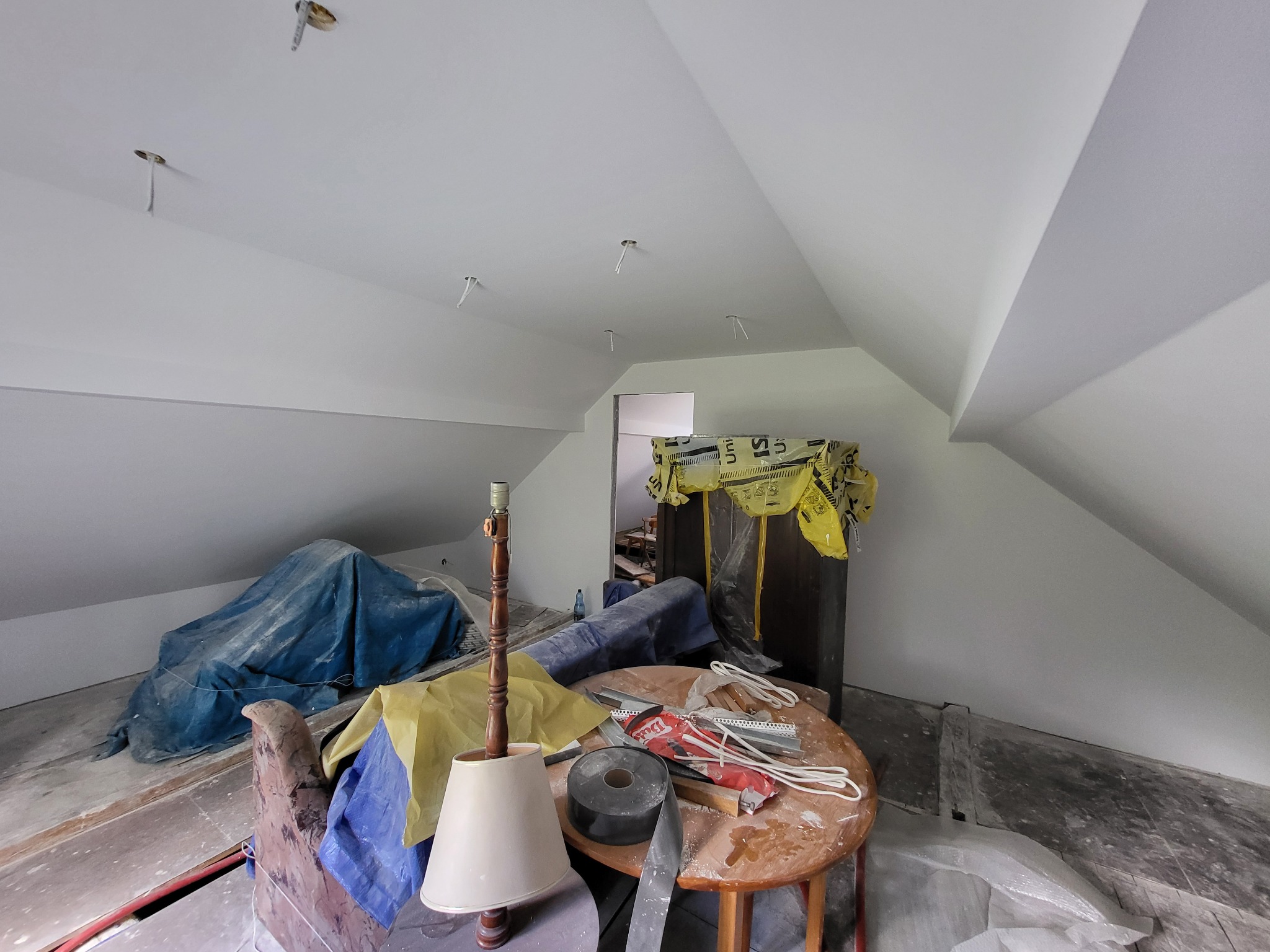 Poddasze po malowaniu na biało, widoczne kable oświetleniowe, meble pod folią i stół z narzędziami. Ściany i sufit świeżo pomalowane, brak lamp.