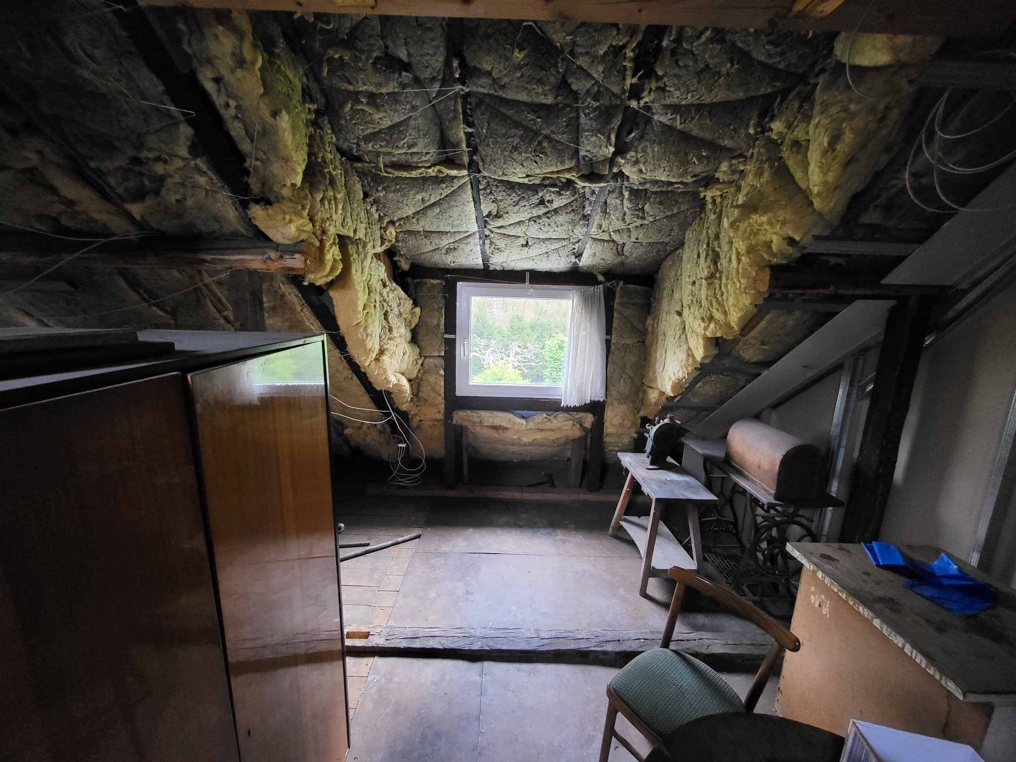 Poddasze z oknem i izolacją ścian wełną mineralną, widoczne stare meble i elementy konstrukcyjne dachu. Ujęcie z perspektywy podłogi, z widocznym oknem w tle.