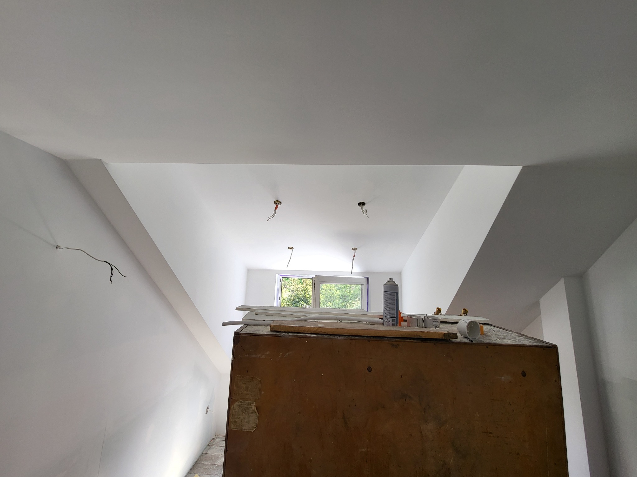 Wnętrze w trakcie prac wykończeniowych: świeżo pomalowany sufit z wystającymi przewodami elektrycznymi, okno w tle, narzędzia i materiały na szafce.