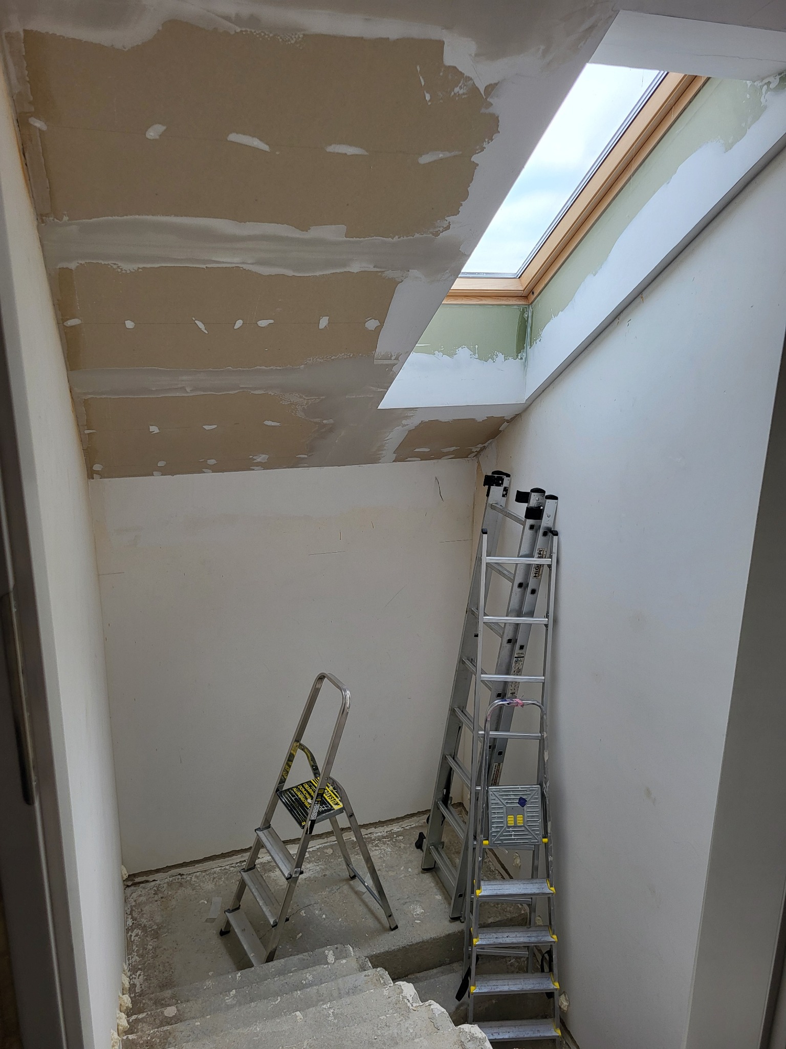 Wnętrze w trakcie prac wykończeniowych: sufit z płyt karton-gips, szpachlowane ściany, okno dachowe. Dwie drabiny opierają się o ścianę, widoczne schody.