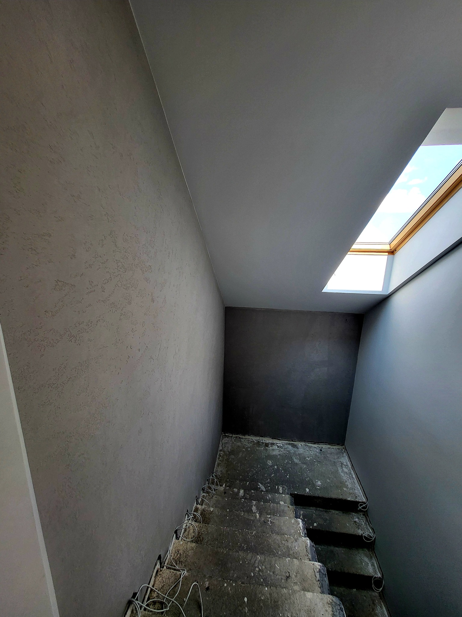 Klatka schodowa w szarościach z oknem dachowym; ściana z fakturą, sufit gładki, schody w stanie surowym z kablami. Widok z dołu, perspektywa zbieżna.