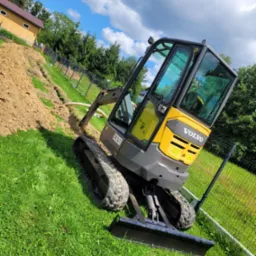 Żółto-szara mini-koparka Volvo ECR25D na trawie, wykonująca wykop pod rurę drenażową wzdłuż ogrodzenia, widok na dom i błękitne niebo z chmurami.