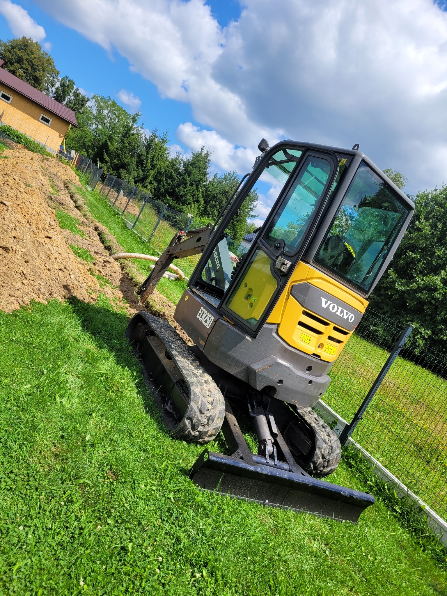 Żółto-szara mini-koparka Volvo ECR25D na trawie, wykonująca wykop pod rurę drenażową wzdłuż ogrodzenia, widok na dom i błękitne niebo z chmurami.