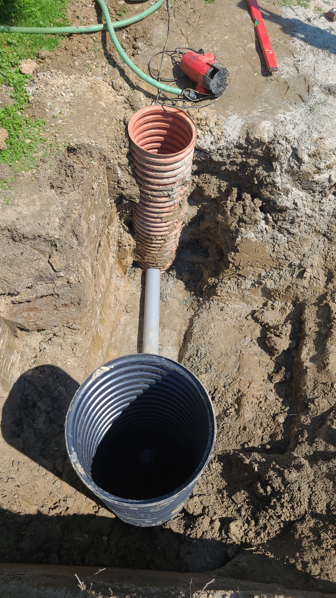 Wykop z widocznymi elementami systemu odwadniającego: perforowaną rurą drenarską w kolorze pomarańczowym, szarą rurą łączącą oraz czarnym zbiornikiem retencyjnym, na górze leżą narzędzia budowlane.