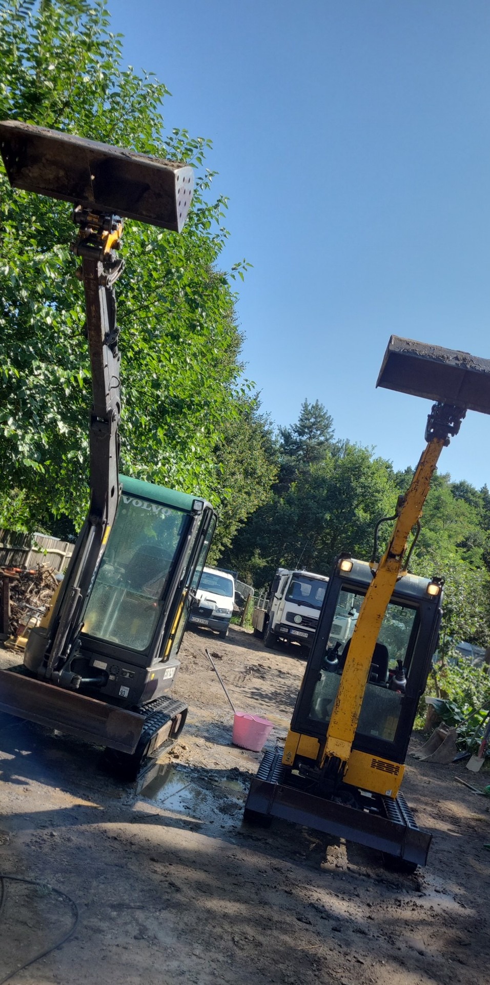 Dwa mini-koparki Volvo i JCB z podniesionymi łyżkami stoją na placu budowy, w tle zaparkowane samochody dostawcze i drzewa.