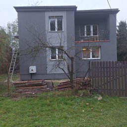 NORBUD - Dwukondygnacyjny dom z szarą elewacją po remoncie, widoczne rusztowanie i stos płyt elewacyjnych przy ogrodzeniu.