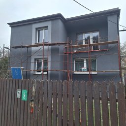 NORBUD - Dwupiętrowy dom w trakcie prac elewacyjnych, otoczony rusztowaniem, widok zza drewnianego płotu z numerem 8 i tabliczką 'Uwaga zły pies'.