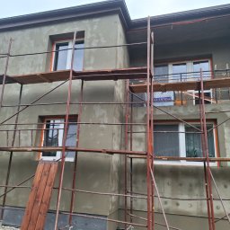 NORBUD - Dom w trakcie ocieplania elewacji, widoczne rusztowania, świeżo nałożony tynk w szarym odcieniu, okna z pomarańczową ramą. Na balkonie widoczne płyty izolacyjne.