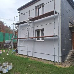 NORBUD - Dom w trakcie prac: część ściany ocieplona szarymi płytami styropianowymi z otworami, druga część pomalowana na szaro, rusztowanie z drewnianymi podestami, na trawie wiadra z zaprawą.