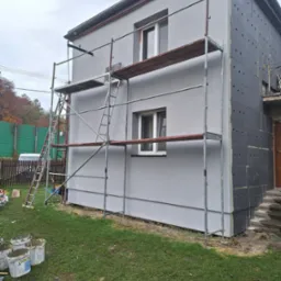 Dom w trakcie prac: część ściany ocieplona szarymi płytami styropianowymi z otworami, druga część pomalowana na szaro, rusztowanie z drewnianymi podestami, na trawie wiadra z zaprawą.