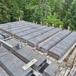 NORBUD - Budowa stropu Teriva z szarych bloczków betonowych i zbrojenia stalowego, widok z góry na tle zieleni drzew.