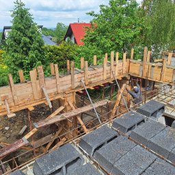 NORBUD - Budowa domu z szalunkiem i betonowymi bloczkami na tle zieleni i domów z czerwoną dachówką, widok z góry na plac budowy.