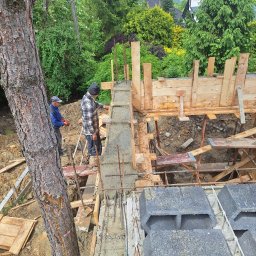 NORBUD - Budowa fundamentów: Dwóch pracowników budowlanych przy pracy, betonowy fundament z drewnianym szalunkiem i stalowym zbrojeniem, widok z góry na plac budowy z drzewem w tle.