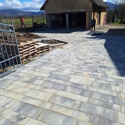 NORBUD - Nowa, szara kostka brukowa ułożona na podjeździe przed budynkiem gospodarczym, palety z materiałem budowlanym i przygotowane podłoże widoczne z boku.