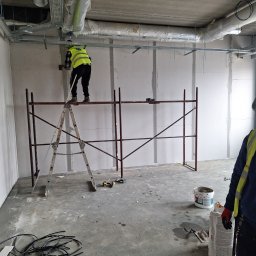 NORBUD - Prace wykończeniowe w surowym wnętrzu: montaż płyt karton-gips na stelażu, pracownik na rusztowaniu w kamizelce odblaskowej, widoczne przewody elektryczne na podłodze i instalacje pod sufitem.