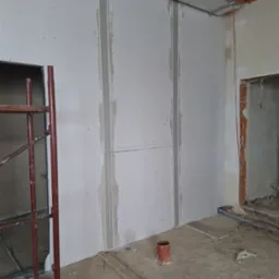 Montaż płyt karton gips na ścianie w trakcie remontu, widoczne szpachlowane łączenia, rusztowanie i elementy instalacji pod sufitem.