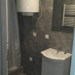 Szara łazienka z bojlerem, umywalką z szafką i grzejnikiem drabinkowym, wykończona płytkami imitującymi marmur.