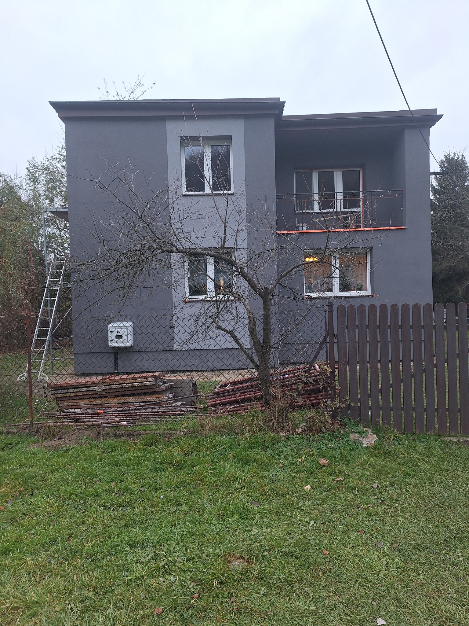 Dwukondygnacyjny dom z szarą elewacją po remoncie, widoczne rusztowanie i stos płyt elewacyjnych przy ogrodzeniu.