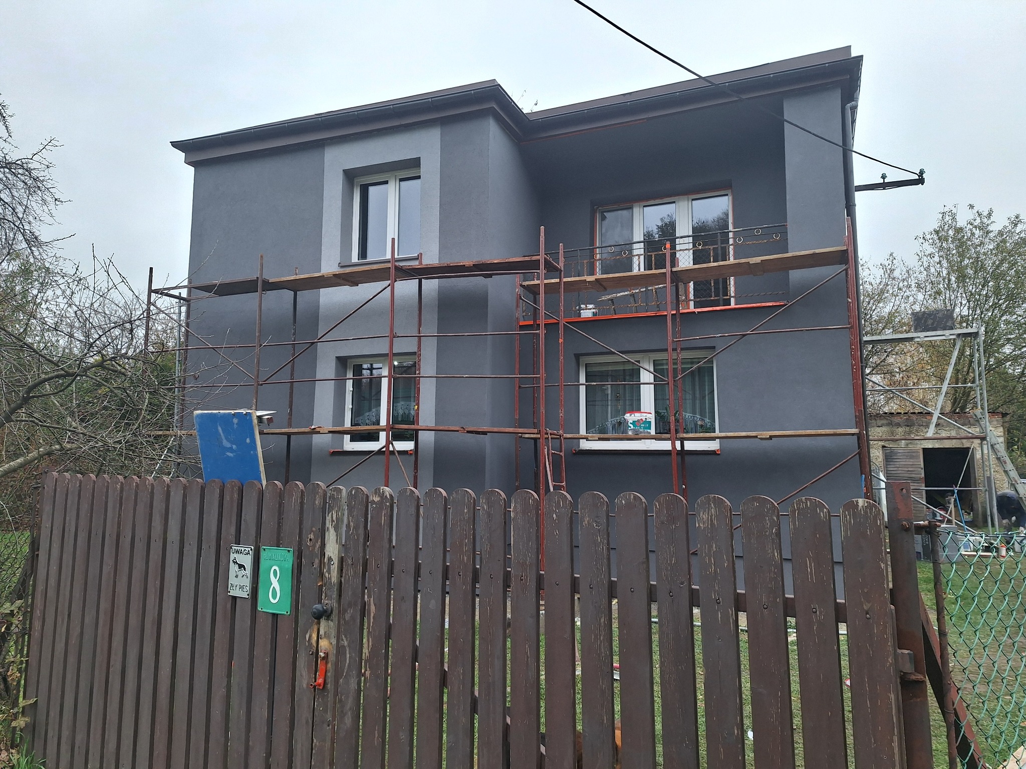 Dwupiętrowy dom w trakcie prac elewacyjnych, otoczony rusztowaniem, widok zza drewnianego płotu z numerem 8 i tabliczką 'Uwaga zły pies'.