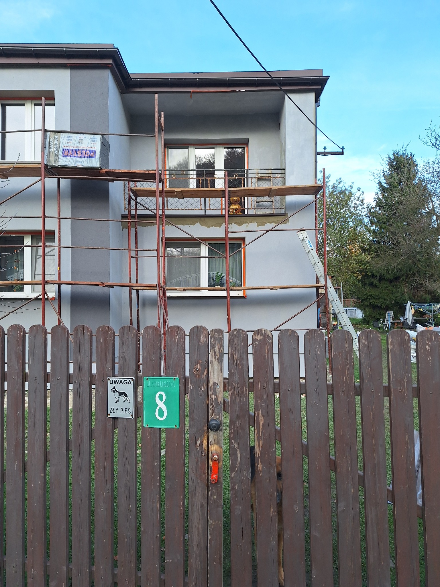 Szary dom jednorodzinny w trakcie prac elewacyjnych, widoczne rusztowania, paczka styropianu na balkonie i drabina oparta o ścianę, brązowy płot z tabliczką z numerem 8 na pierwszym planie, pies...
