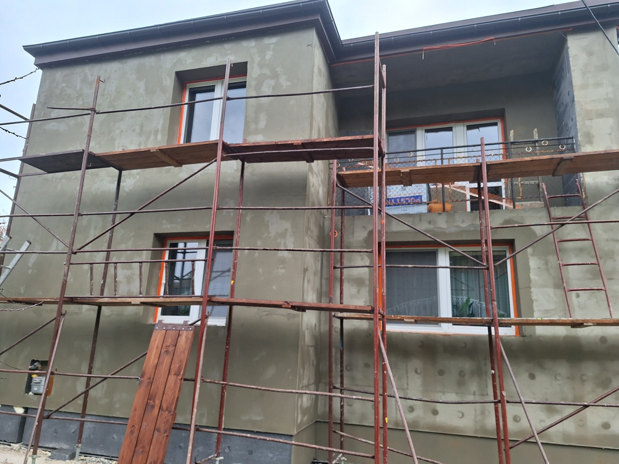 Dom w trakcie ocieplania elewacji, widoczne rusztowania, świeżo nałożony tynk w szarym odcieniu, okna z pomarańczową ramą. Na balkonie widoczne płyty izolacyjne.