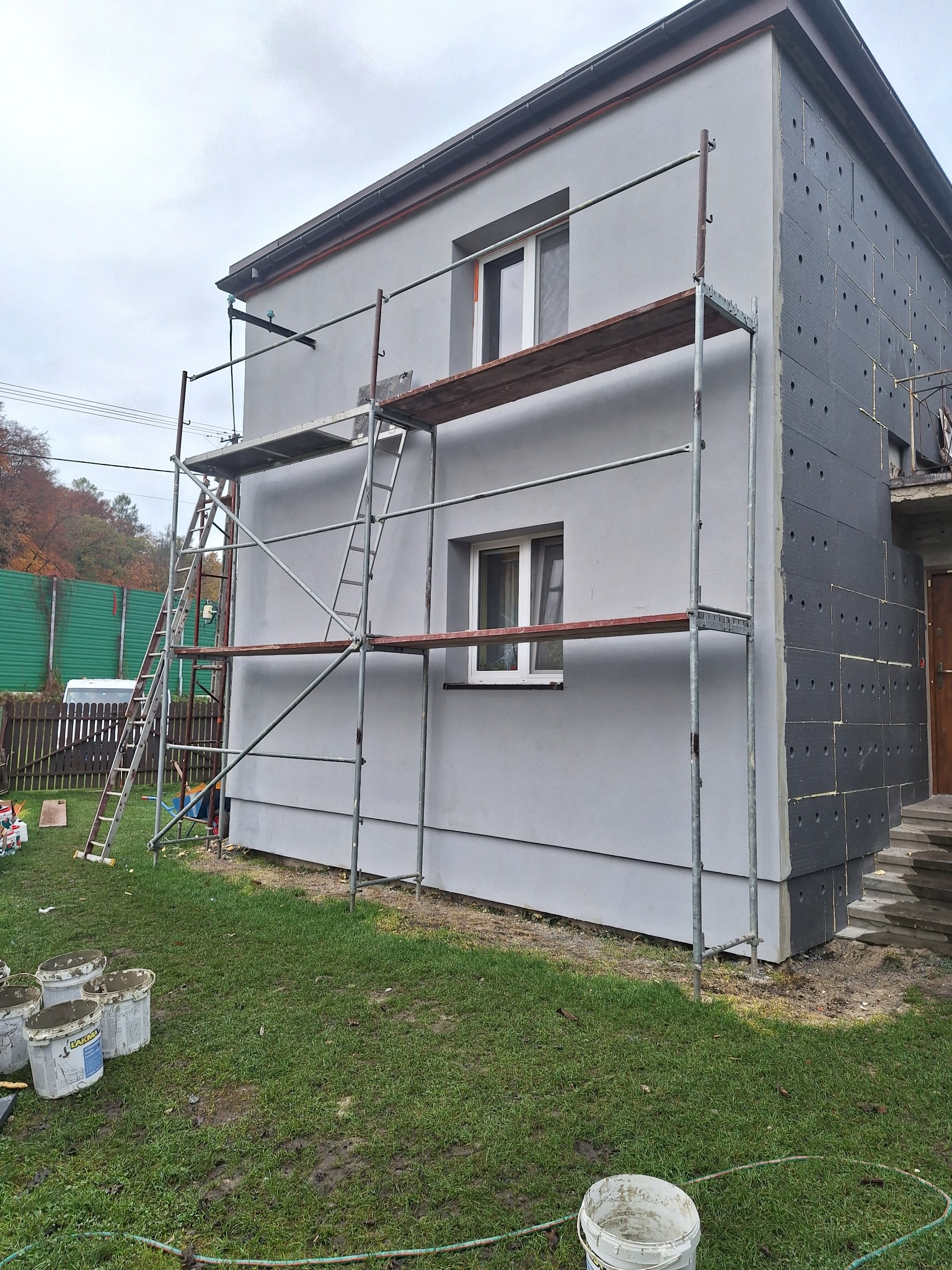 Dom w trakcie prac: część ściany ocieplona szarymi płytami styropianowymi z otworami, druga część pomalowana na szaro, rusztowanie z drewnianymi podestami, na trawie wiadra z zaprawą.