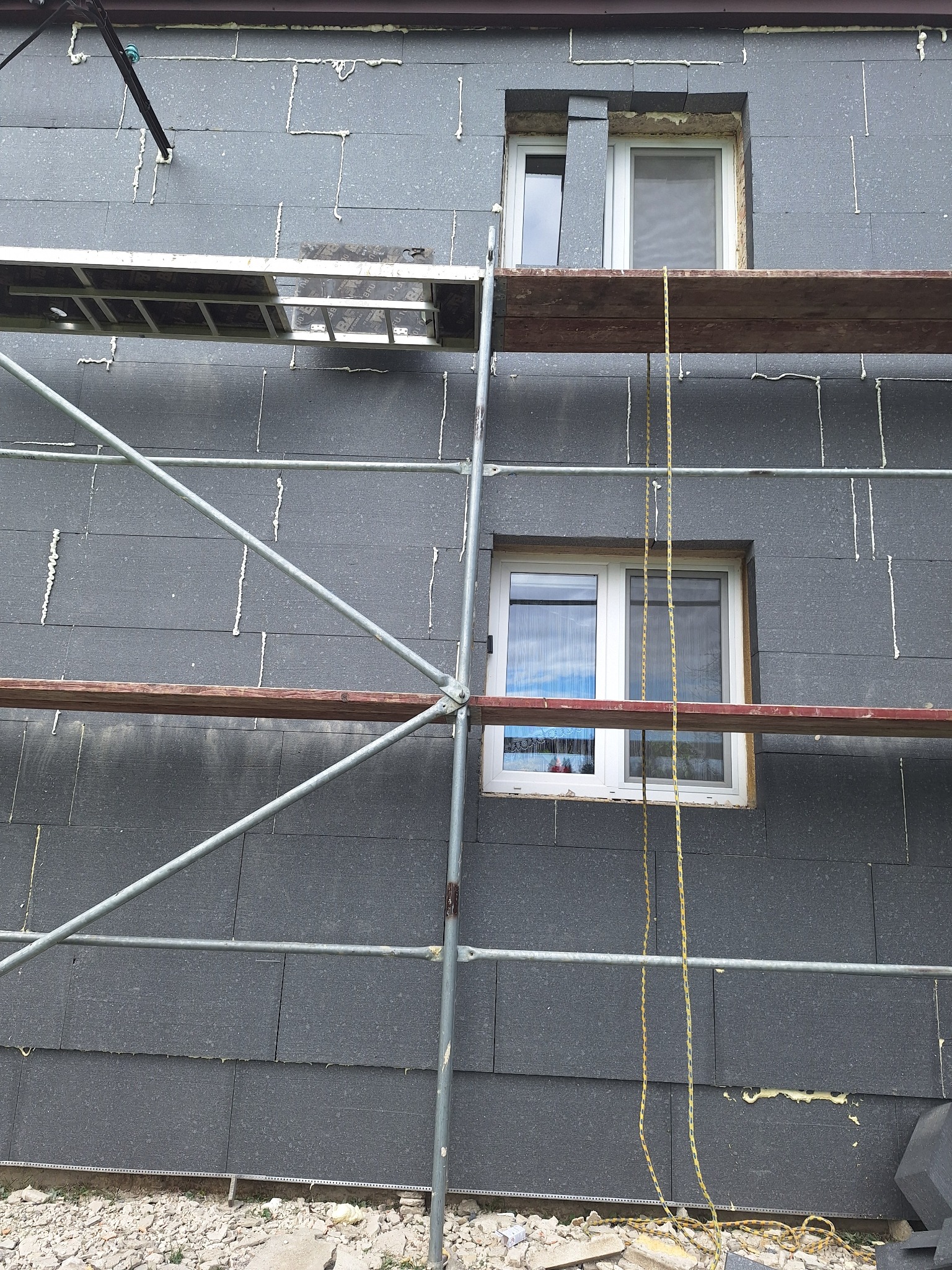 Ściana budynku pokryta szarym styropianem, widoczne rusztowanie i szczeliny wypełnione pianką montażową, przygotowanie do dalszych prac wykończeniowych.