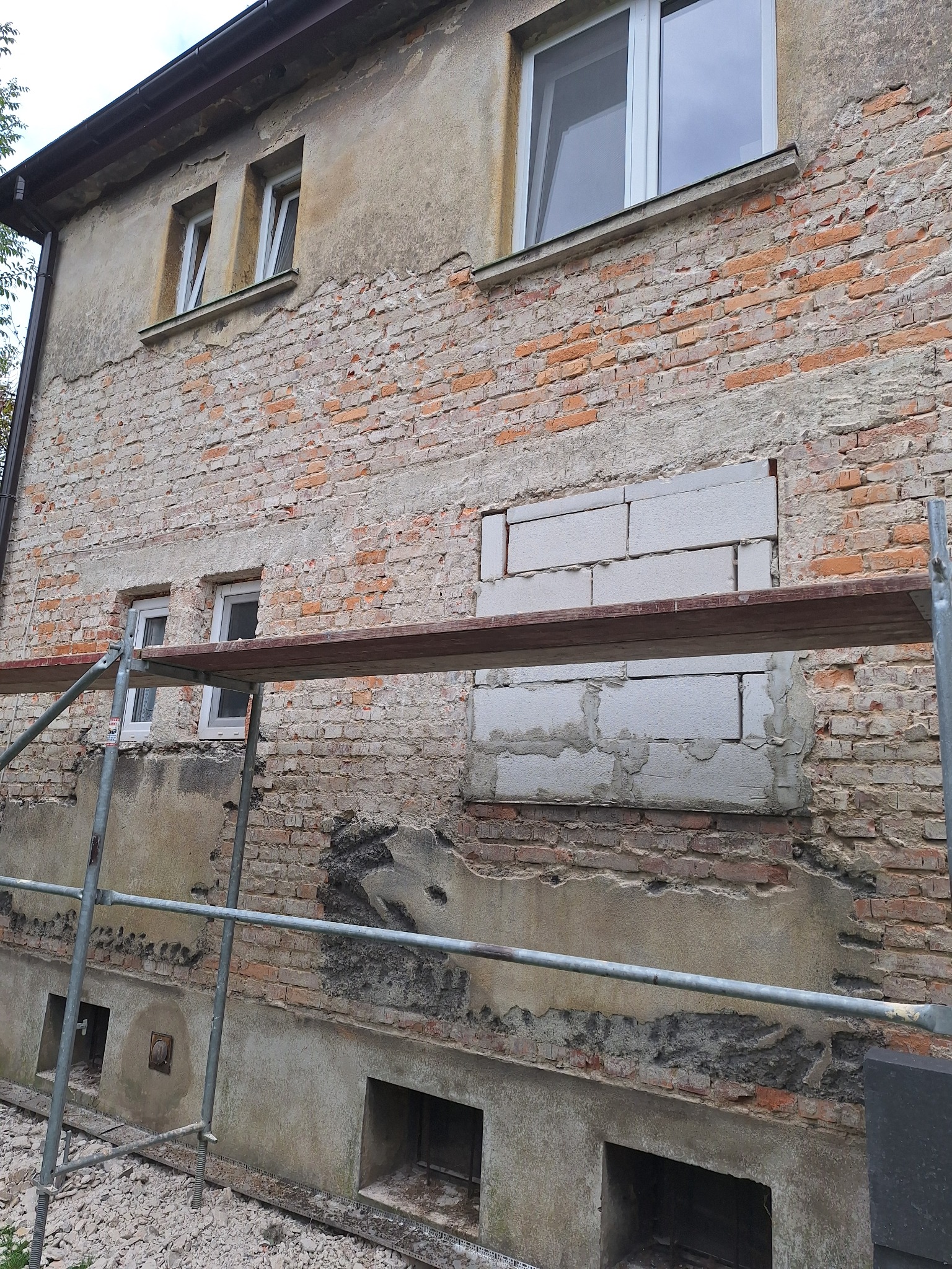 Niedokończona elewacja budynku z widocznym murem z czerwonej cegły, fragmentami starego tynku, nowymi oknami PCV i zamurowanymi otworami z szarego betonu komórkowego, częściowo zasłonięta...