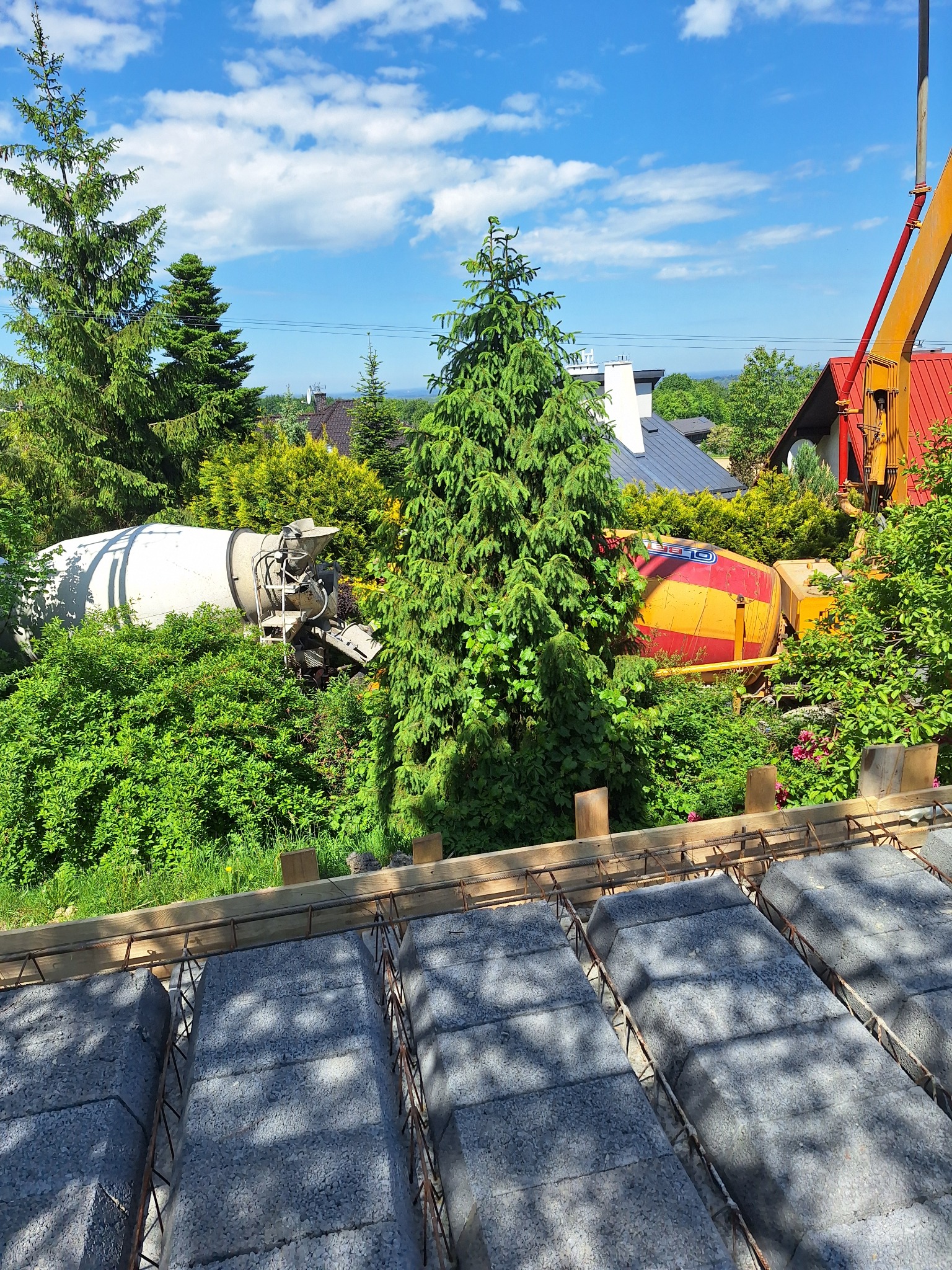 Widok z góry na przygotowany szalunek stropu Teriva z widocznymi betonowymi pustakami i zbrojeniem, w tle dwa betoniarki ukryte w zieleni i żuraw budowlany.