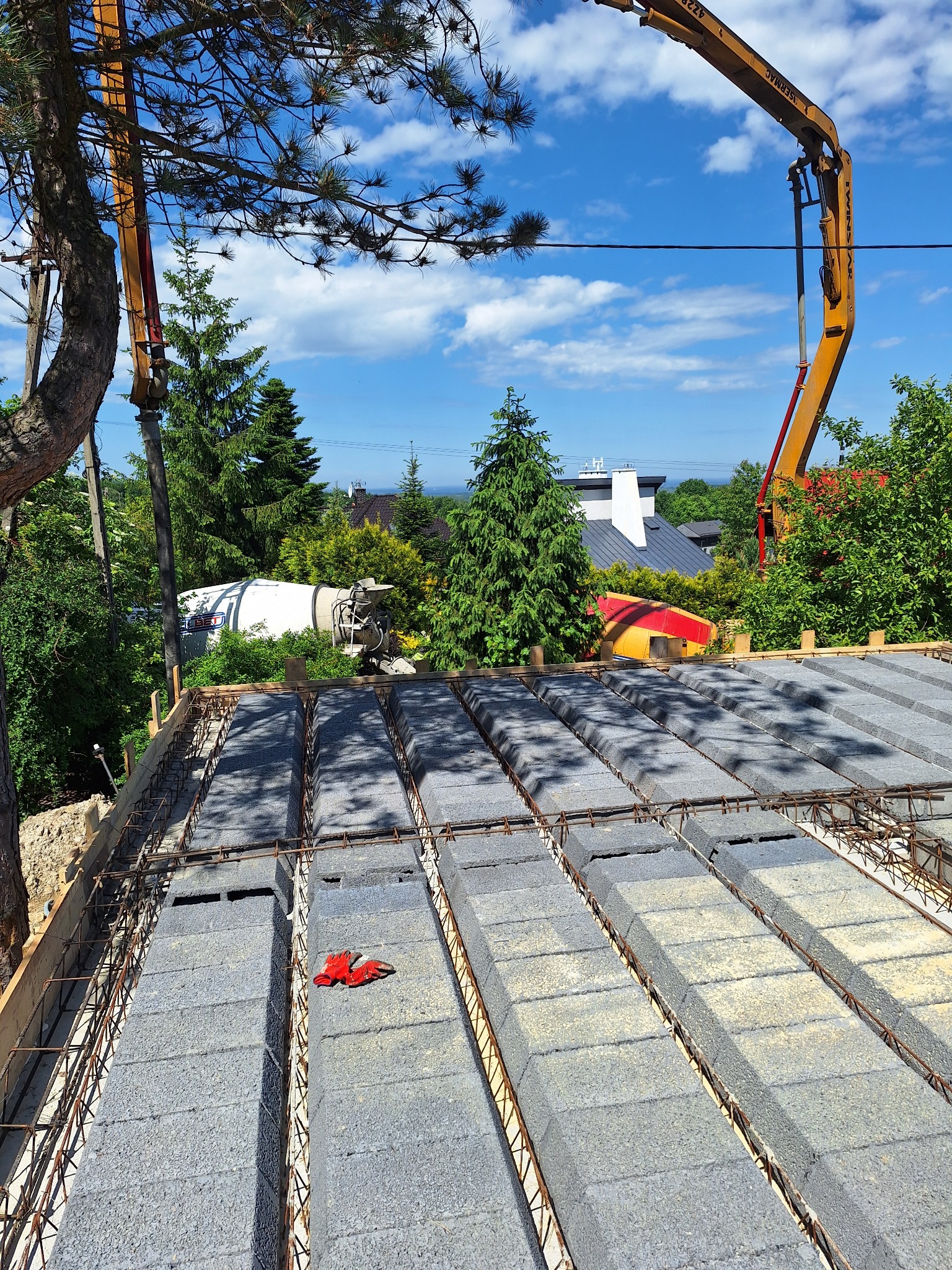 Wiązanie zbrojenia na stropie Teriva z pustaków szalunkowych, widoczna pompa do betonu i betonomieszarka w tle, na pustakach leżą czerwone rękawice robocze.