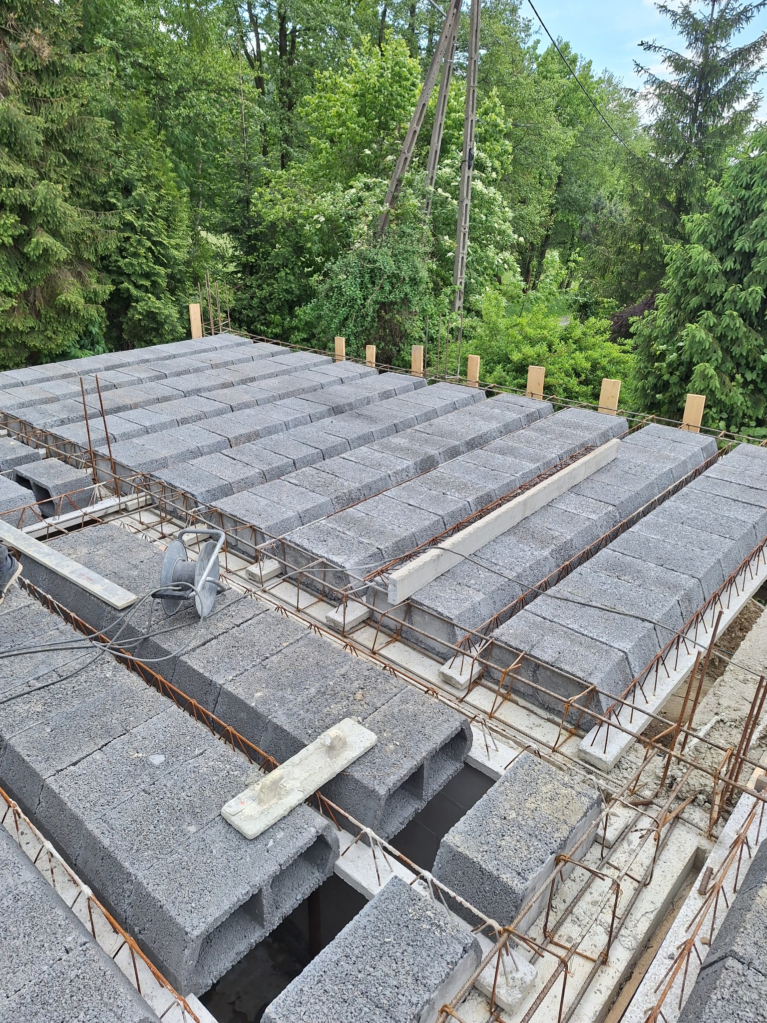 Budowa stropu Teriva z szarych bloczków betonowych i zbrojenia stalowego, widok z góry na tle zieleni drzew.
