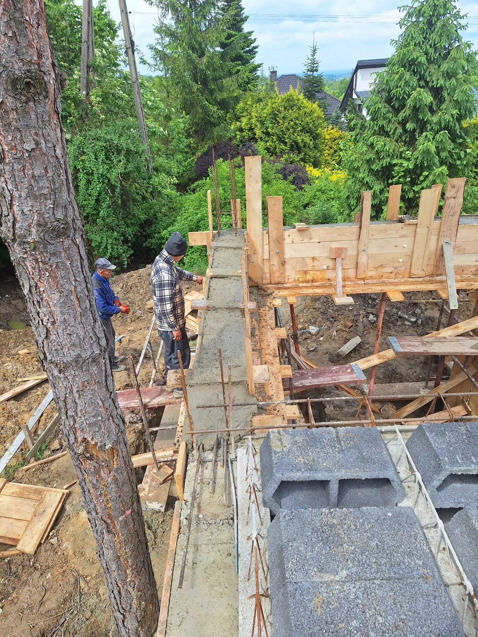 Budowa fundamentów: Dwóch pracowników budowlanych przy pracy, betonowy fundament z drewnianym szalunkiem i stalowym zbrojeniem, widok z góry na plac budowy z drzewem w tle.