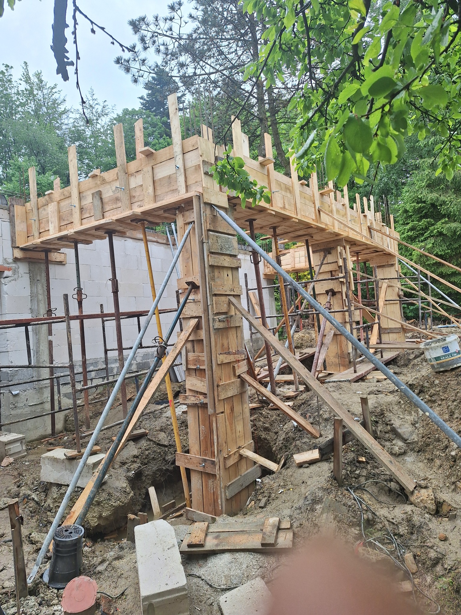 Szare bloczki fundamentowe z drewnianym szalunkiem wzmocnionym metalowymi podporami, widok na plac budowy w pochmurny dzień.