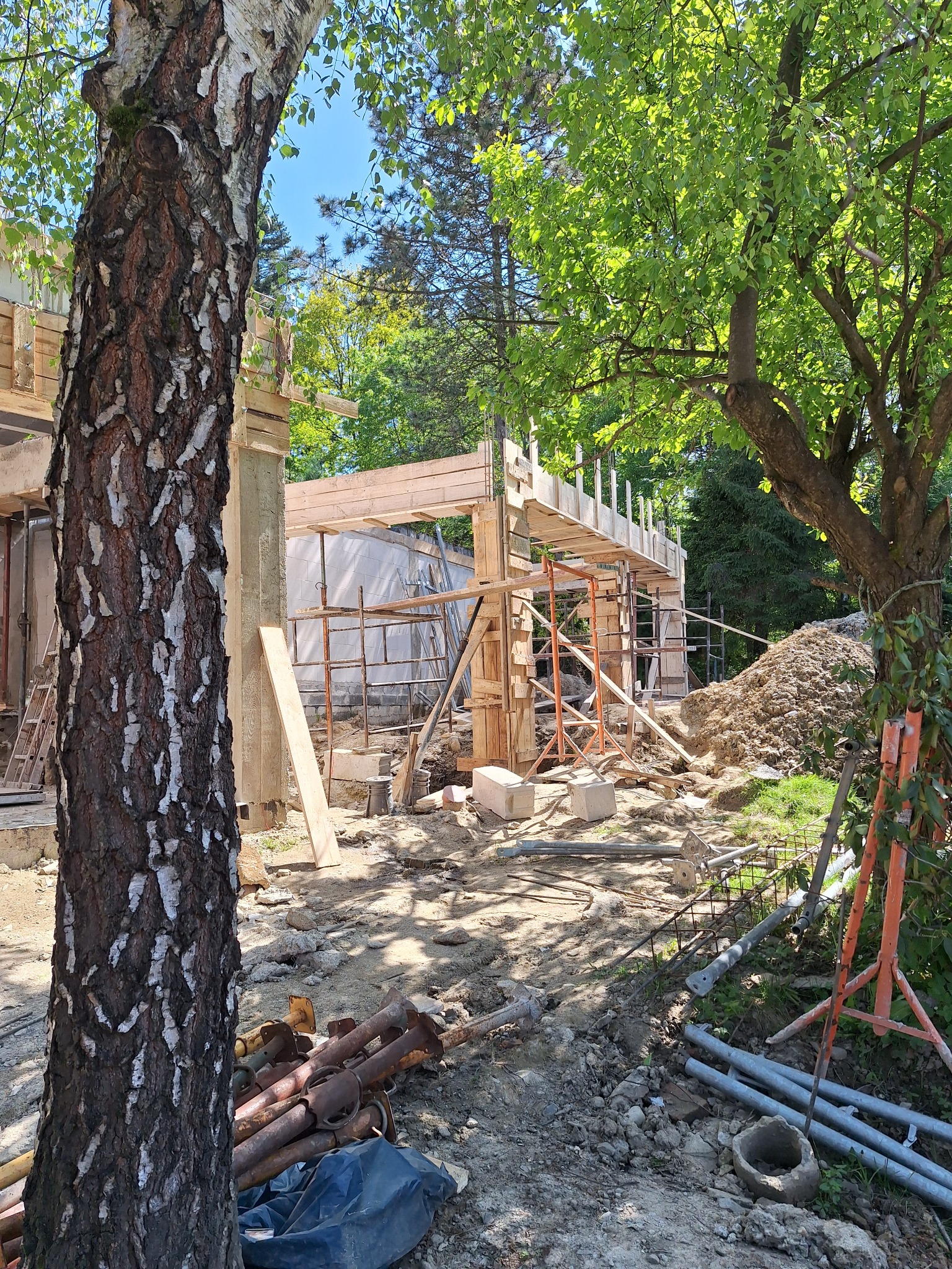 Budowa domu w trakcie realizacji, widoczne drewniane szalunki, rusztowania i stos ziemi, z przodu brzoza.