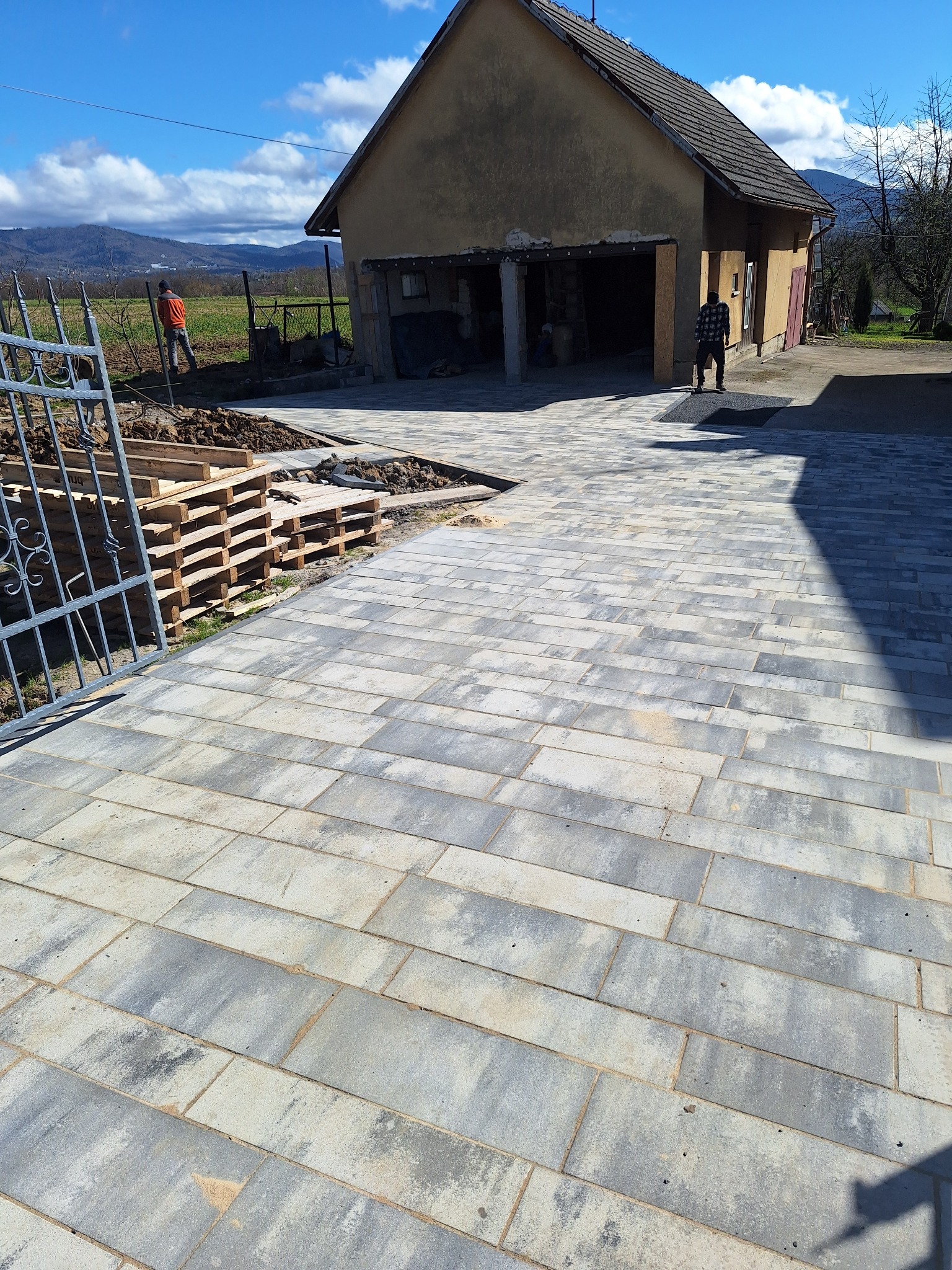 Nowa, szara kostka brukowa ułożona na podjeździe przed budynkiem gospodarczym, palety z materiałem budowlanym i przygotowane podłoże widoczne z boku.