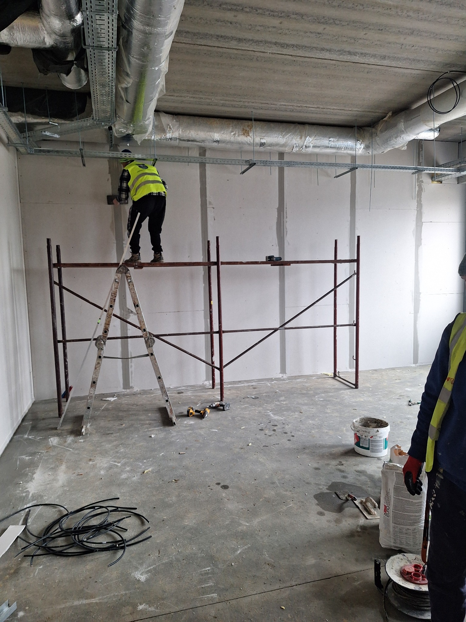 Prace wykończeniowe w surowym wnętrzu: montaż płyt karton-gips na stelażu, pracownik na rusztowaniu w kamizelce odblaskowej, widoczne przewody elektryczne na podłodze i instalacje pod sufitem.