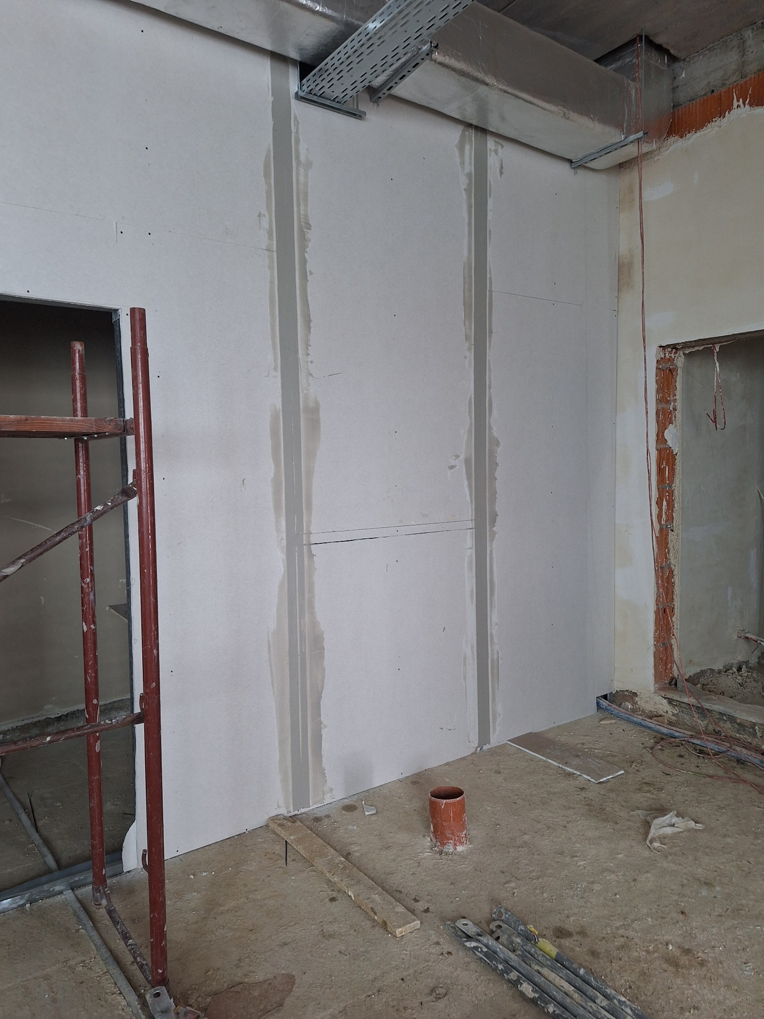 Montaż płyt karton gips na ścianie w trakcie remontu, widoczne szpachlowane łączenia, rusztowanie i elementy instalacji pod sufitem.