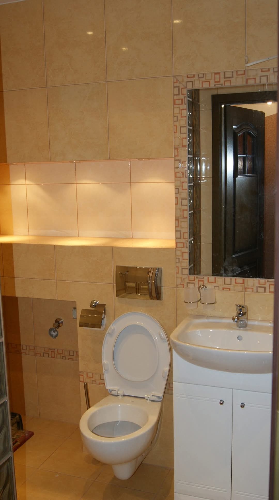 Wykończona łazienka z beżowymi płytkami, podświetlaną półką, lustrem z dekoracyjną ramą i wiszącą toaletą z otwartą deską.