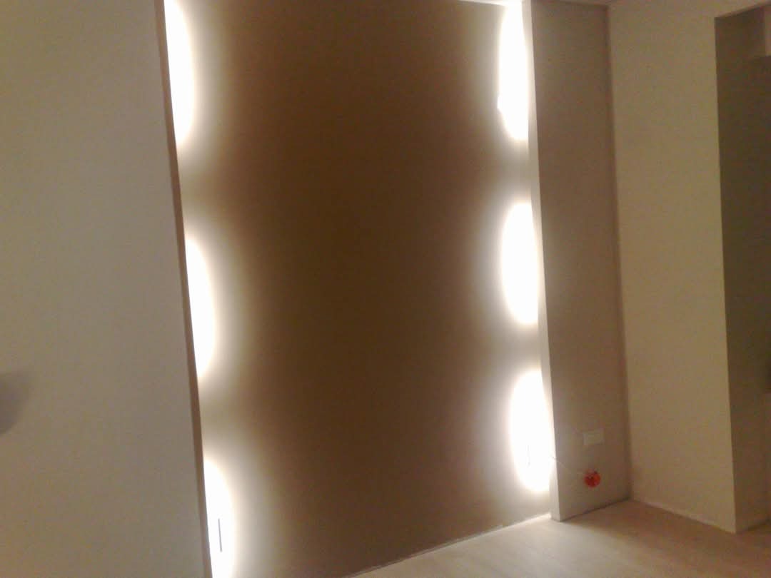 Beżowa ściana z pionowymi wnękami oświetlonymi ledowym światłem, widoczny fragment parkietu i gniazdko elektryczne.