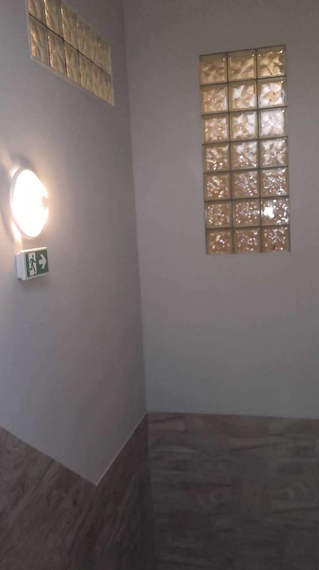 Korytarz z oświetleniem awaryjnym i dwoma oknami z luksferów, wykończony płytkami imitującymi drewno.