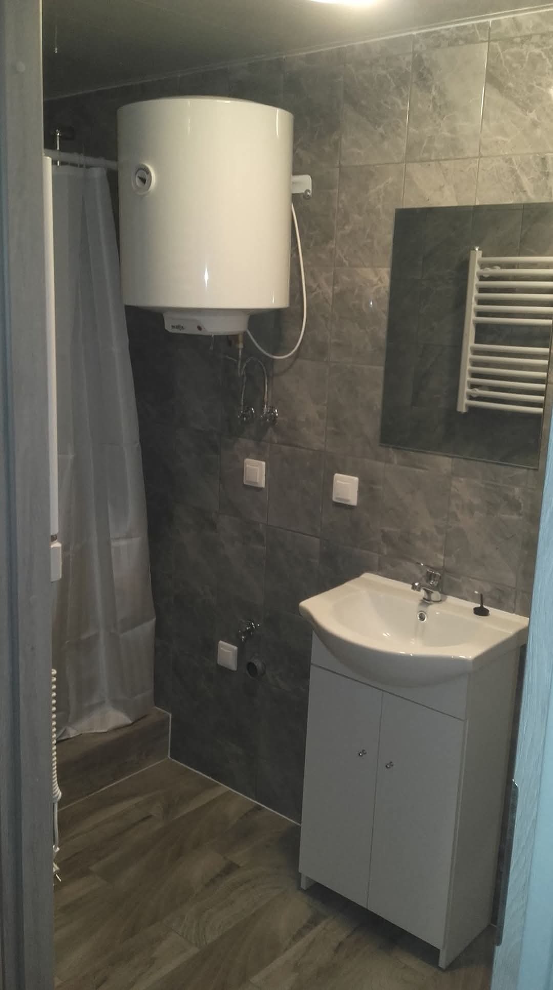 Szara łazienka z bojlerem, umywalką z szafką i grzejnikiem drabinkowym, wykończona płytkami imitującymi marmur.