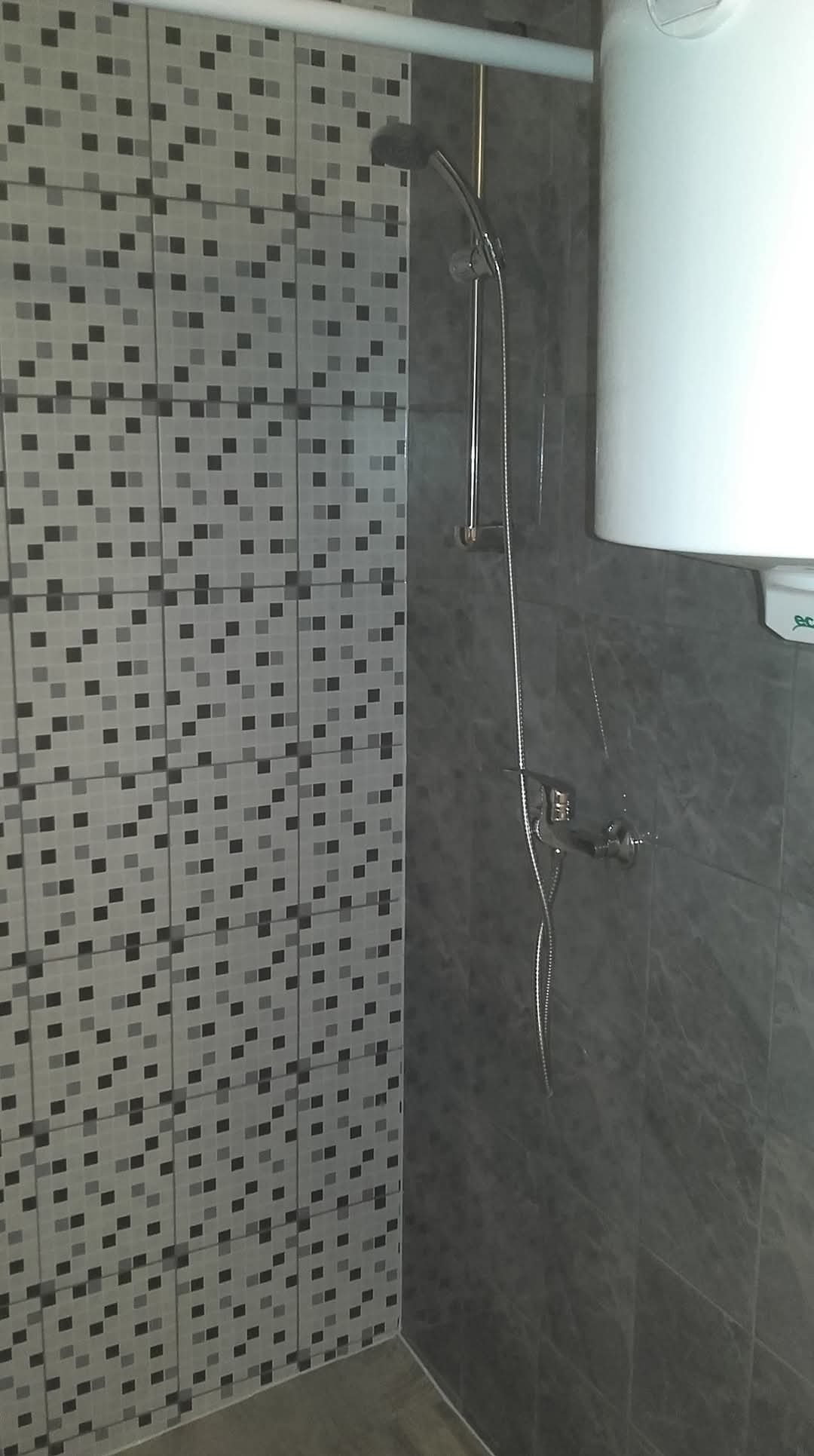 Wykończona łazienka z szaro-białą mozaikową glazurą, chromowaną armaturą prysznicową i białym bojlerem na ścianie z ciemnoszarych płytek.