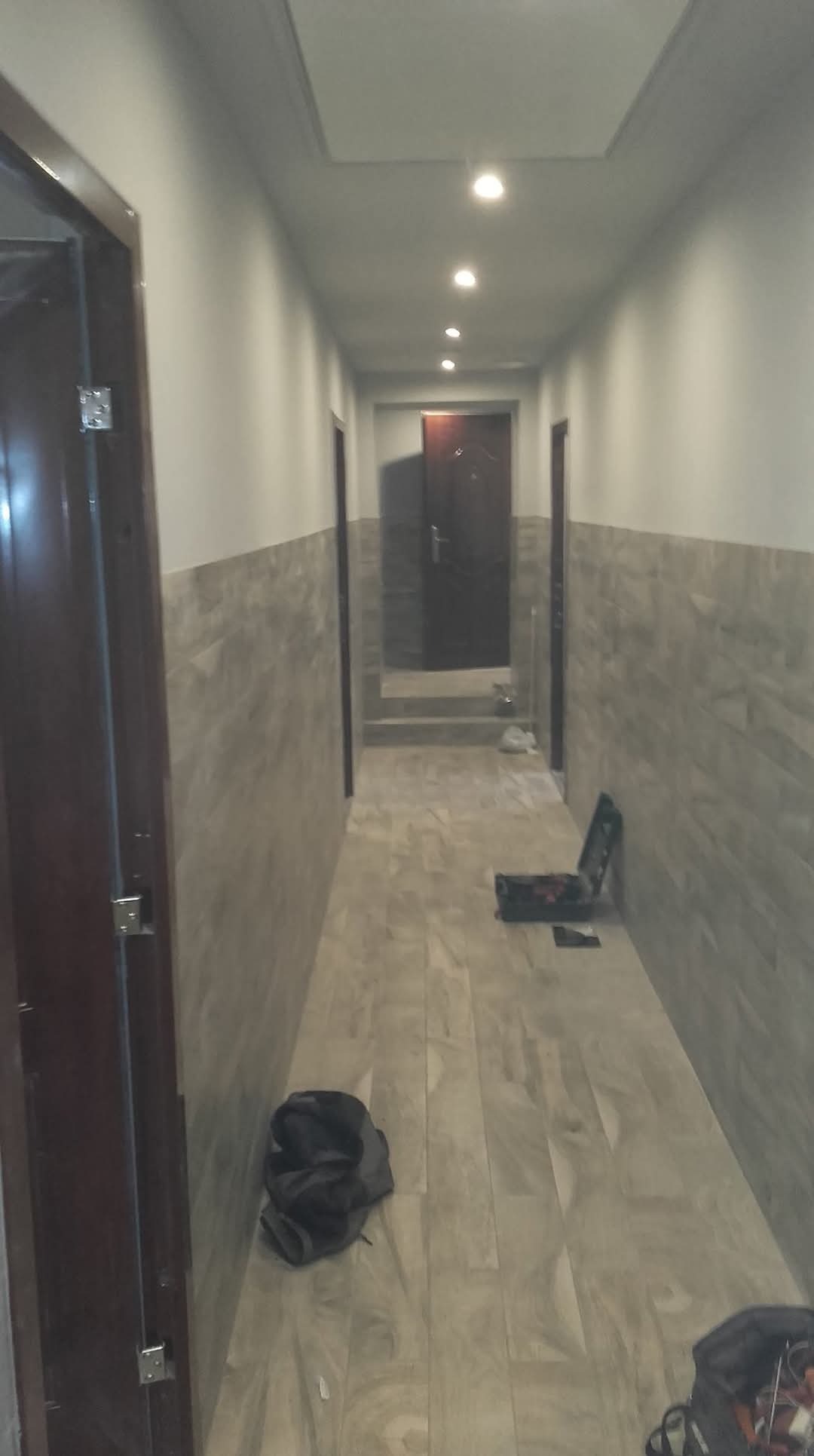 Długi korytarz w trakcie prac wykończeniowych, z położoną częściowo płytką imitującą drewno na ścianach i podłodze, widoczne narzędzia i torby robocze.
