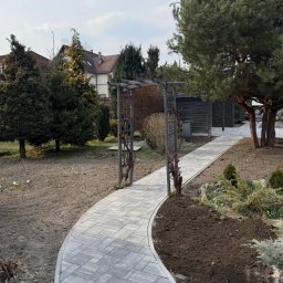 Oleksandr Chyplyk - Ścieżka z szarej kostki brukowej wijąca się przez ogród, z metalową pergolą i świeżo obsadzoną rabatą. W tle domy i drzewa.