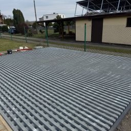 Oleksandr Chyplyk - Ułożona szara kostka ażurowa na posesji, widoczne narzędzia brukarskie i budynek gospodarczy z panelami słonecznymi w tle. Kąt ujęcia z dołu podkreśla regularność wzoru.