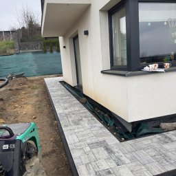 Oleksandr Chyplyk - Ułożona ścieżka z szarej kostki brukowej przy białym budynku. Widoczna zagęszczarka, ziemia i fragment zielonego ogrodzenia. Prace brukarskie w toku.