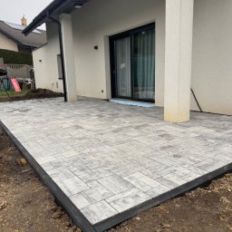 Oleksandr Chyplyk - Nowoczesny taras z jasnoszarej kostki brukowej ułożonej przy domu z dużym oknem przesuwnym. Widoczne obrzeża i przygotowane podłoże.