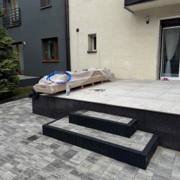 Oleksandr Chyplyk - Ułożona kostka brukowa w szarym odcieniu prowadząca do schodów z czarnymi obrzeżami i tarasu. Na tarasie zapakowane elementy, butelka wody i niebieski wąż.