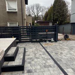 Oleksandr Chyplyk - Nowoczesna, szara kostka brukowa z czarnymi obrzeżami i schodami, brama w tle. Widoczna ubijarka i narzędzia. Realizacja z drenażem liniowym.